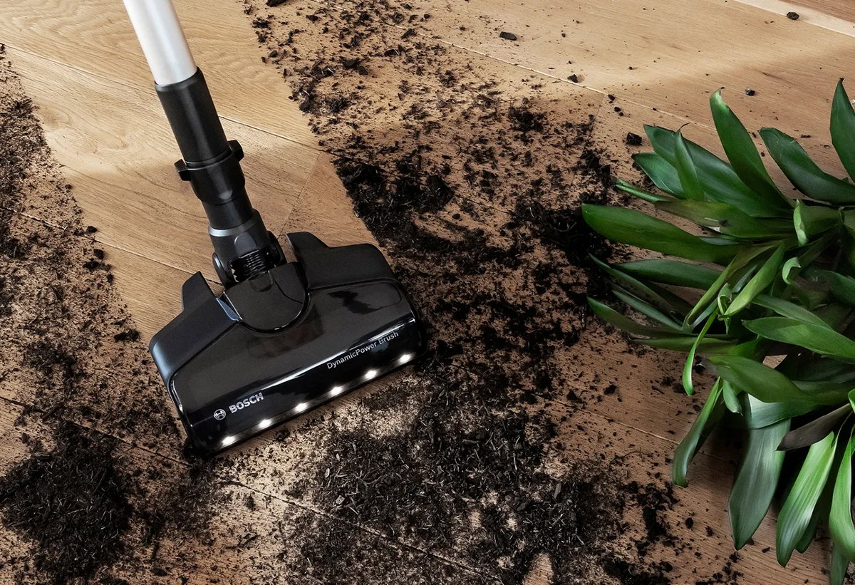 Σκούπα Bosch με βούρτσα DynamicPower Brush που καθαρίζει σκόνη και χώμα από ξύλινο πάτωμα, δίπλα σε πράσινο φυτό